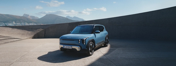 Frontansicht des blauen Kia EV2 auf einem modernen Vorplatz vor Bergkulisse.